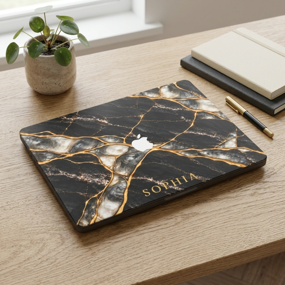 Top view of a MacBook in a Black Pearl marble case personalized with SOPHIA, Apple logo visible, on a wooden desk with a plant.