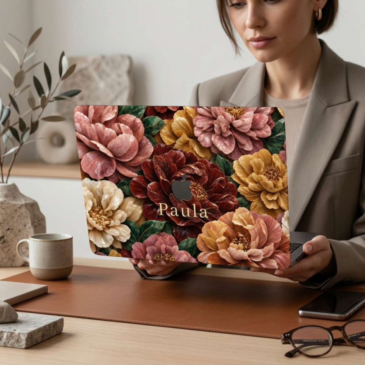Personalized TrioAuraDesign MacBook case featuring a Stone Bouquet design with blush pink, burgundy, ivory and warm golden stone-effect flowers, deep green leaves and the custom name Paula. A woman in a neutral blazer holds the laptop at a modern desk with a coffee cup, phone, glasses, stone decor and soft botanical accents, with the Apple logo visible in the center.