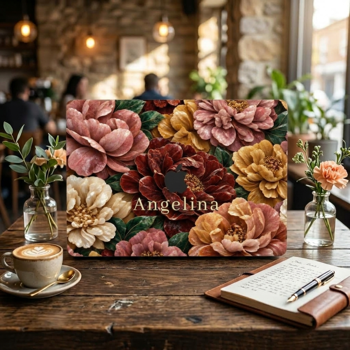 Personalized TrioAuraDesign MacBook case featuring a Stone Bouquet design with blush pink, burgundy, ivory and warm golden stone-effect flowers, deep green leaves and the custom name Angelina. The MacBook is displayed upright on a rustic wooden café table beside a cappuccino, open notebook, pen and small flower vases, with warm pendant lights and a softly blurred café interior in the background.