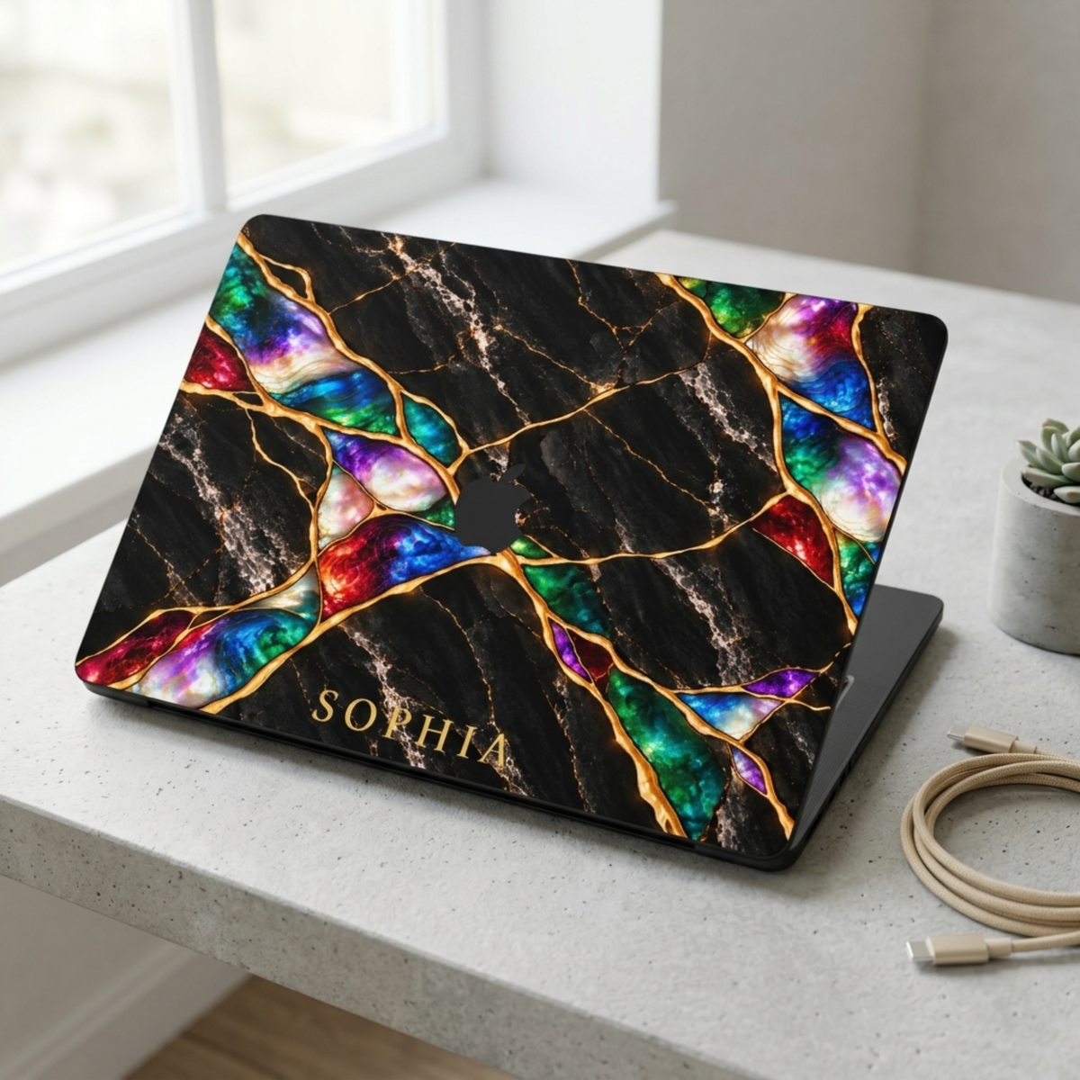 MacBook in a Jewel Tone black marble hard shell case with gold veins and colorful opal inlay, personalized with SOPHIA, on a white desk. Printed effect, smooth matte finish.
