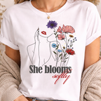 Close-up portrait of a woman wearing a white "She Blooms Softly" T-shirt, highlighting the minimalist line art of a woman's face integrated with colorful wildflowers.