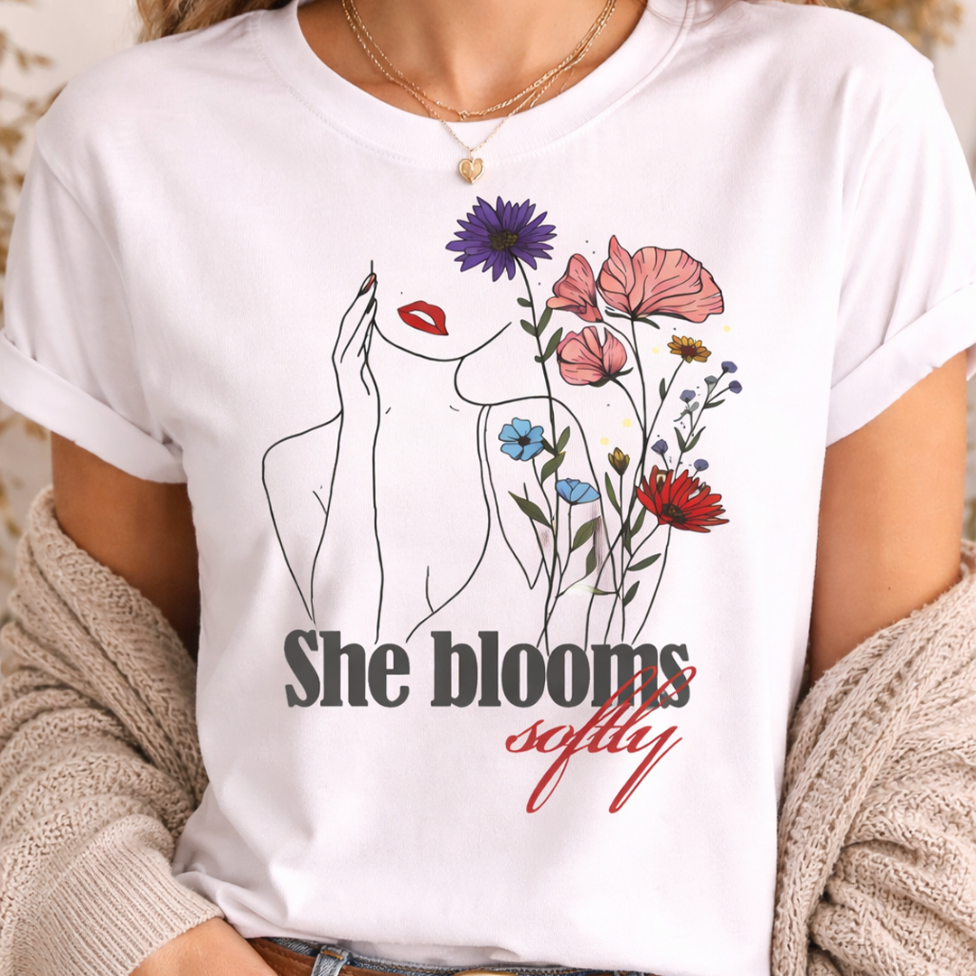 Close-up portrait of a woman wearing a white "She Blooms Softly" T-shirt, highlighting the minimalist line art of a woman's face integrated with colorful wildflowers.