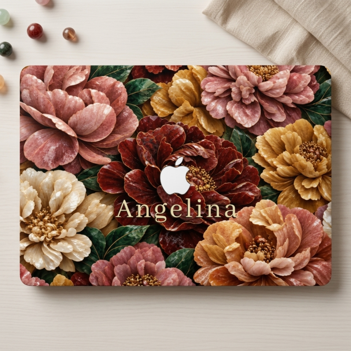Personalized TrioAuraDesign MacBook case featuring a Stone Bouquet design with large blush pink, burgundy, ivory and warm golden flowers, deep green leaves and the custom name Angelina. The Apple logo is visible in the center, with the case styled on a light neutral desk beside beige fabric and small decorative stones.