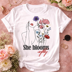 Flatlay mockup of a white "She Blooms Softly" cotton tee with vibrant wildflowers and line art design, styled with fresh pink peonies and aesthetic decor.