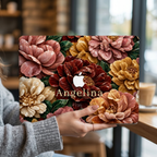 Personalized TrioAuraDesign MacBook case with a Stone Bouquet design featuring blush pink, burgundy, ivory and warm golden stone-effect flowers, deep green leaves and the custom name Angelina. The laptop is held upright in a cozy café setting, with the Apple logo visible in the center and a softly blurred window and coffee cup in the background.
