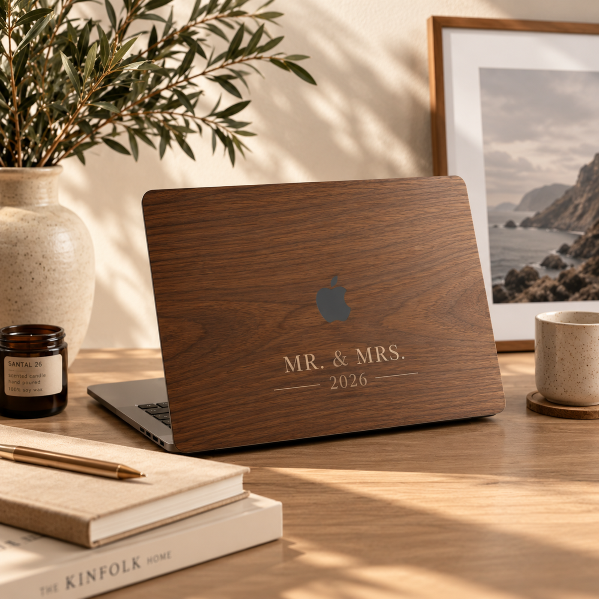Personalized wood grain effect MacBook case by TrioAuraDesign with the custom text MR. & MRS. 2026, shown on a laptop on a sunlit wooden desk with olive branches in a ceramic vase, a candle, books and a framed coastal print. The dark walnut wood look and lettering are UV-printed on a smooth polycarbonate hard shell, not real wood or engraving. A personalized wedding, anniversary or Christmas gift for a newly married couple.