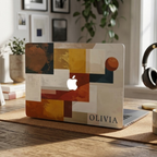 Personalized abstract geometric MacBook case with custom name Olivia in terracotta mustard navy and cream color blocks, displayed on rustic wooden desk with headphones, books, plant and framed art gallery wall in background — bold architectural collage hard shell laptop cover with elegant serif name personalization, Apple logo visible through an unprinted area, slim snap-on fit, for MacBook Air 13 M2 M3 M4 M5 and MacBook Pro 14 16 M3 M4 M5, modern aesthetic personalized gift for creative professionals