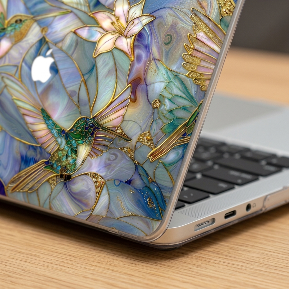 Close-up detail of a stained glass hummingbird MacBook case by TrioAuraDesign on a partially open laptop. The image highlights the slim hard-shell profile, precise port access, and smooth protective finish. The artwork features a vibrant hummingbird, white lilies, and flowing blue, teal, lavender, and pearl stained glass patterns with metallic gold outlines. The visible Apple logo blends naturally into the design, showcasing the premium print quality for MacBook Air and MacBook Pro models.