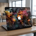 Front lifestyle photo of a personalized artistic world map MacBook case by TrioAuraDesign displayed on a partially open laptop. The hard-shell case features a colorful abstract world map with vibrant watercolor textures, dark wood background details, and the personalized name "Kristen" in elegant white script. The image highlights the smooth protective finish, slim lightweight profile, unique travel-inspired artwork, and precise fit for MacBook Air and MacBook Pro models.