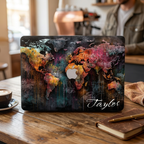 Lifestyle photo of a personalized artistic world map MacBook case by TrioAuraDesign displayed on a partially open laptop in a cozy café. The hard-shell case features a colorful abstract world map with vibrant watercolor textures, dark wood background details, and the personalized name "Taylor" in elegant white script. The image highlights the smooth protective finish, slim lightweight profile, bold travel-inspired artwork, and custom fit for MacBook Air and MacBook Pro models.