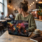 Lifestyle photo of a personalized artistic world map MacBook case by TrioAuraDesign displayed on a partially open laptop in a cozy café setting. The hard-shell case features a colorful abstract world map with vibrant watercolor textures, dark wood background details, and the personalized name "Kristen" in elegant white script. The image highlights the smooth protective finish, slim lightweight profile, unique travel-inspired artwork, and custom fit for MacBook Air and MacBook Pro models.
