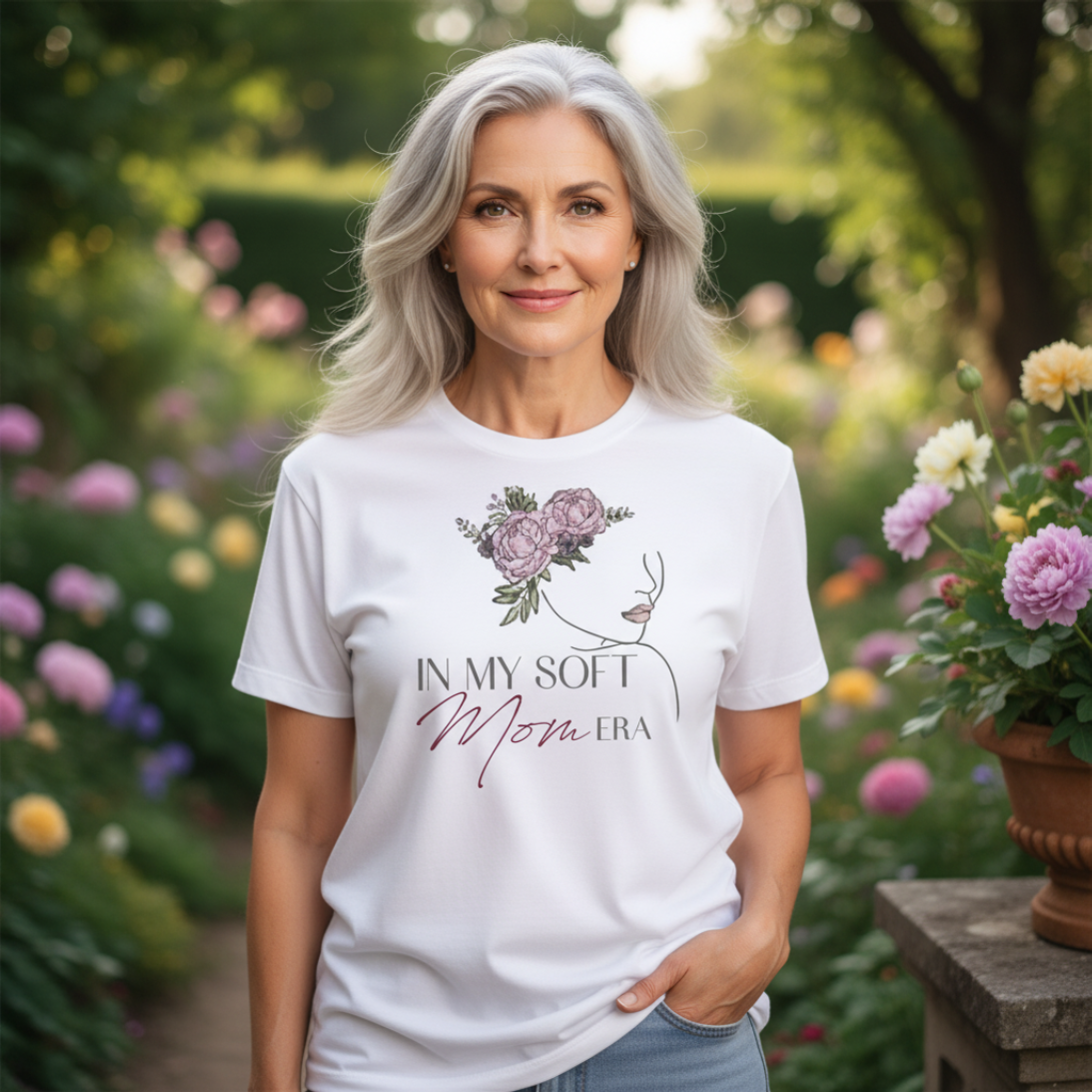 Happy older woman with grey hair wearing a white "In My Soft Mom Era" T-shirt, smiling while posing in a vibrant flower garden.