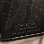 Close-up of a personalized City Map MacBook case from TrioAuraDesign, featuring a dark New York street map with custom city text “NEW YORK,” coordinates, and the phrase “YOUR PLACE. YOUR STORY.” The detailed map lines and warm beige lettering are shown up close on the smooth printed hard shell, highlighting the custom map design and personalization details.