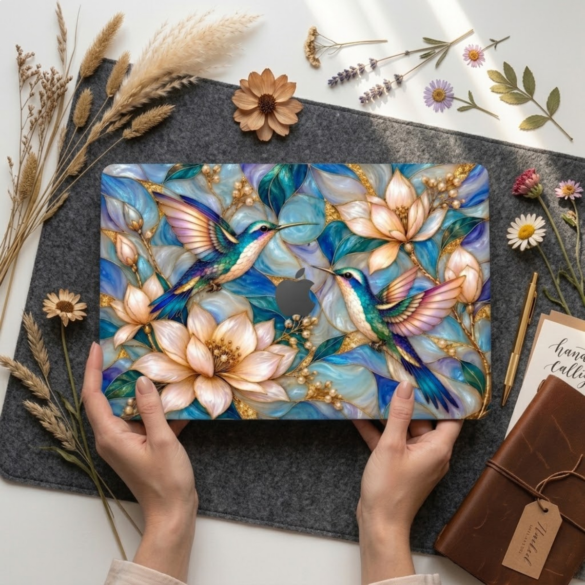 Hands placing a MacBook in the hummingbird magnolia stained glass effect case on a grey felt mat surrounded by dried flowers and a leather notebook.