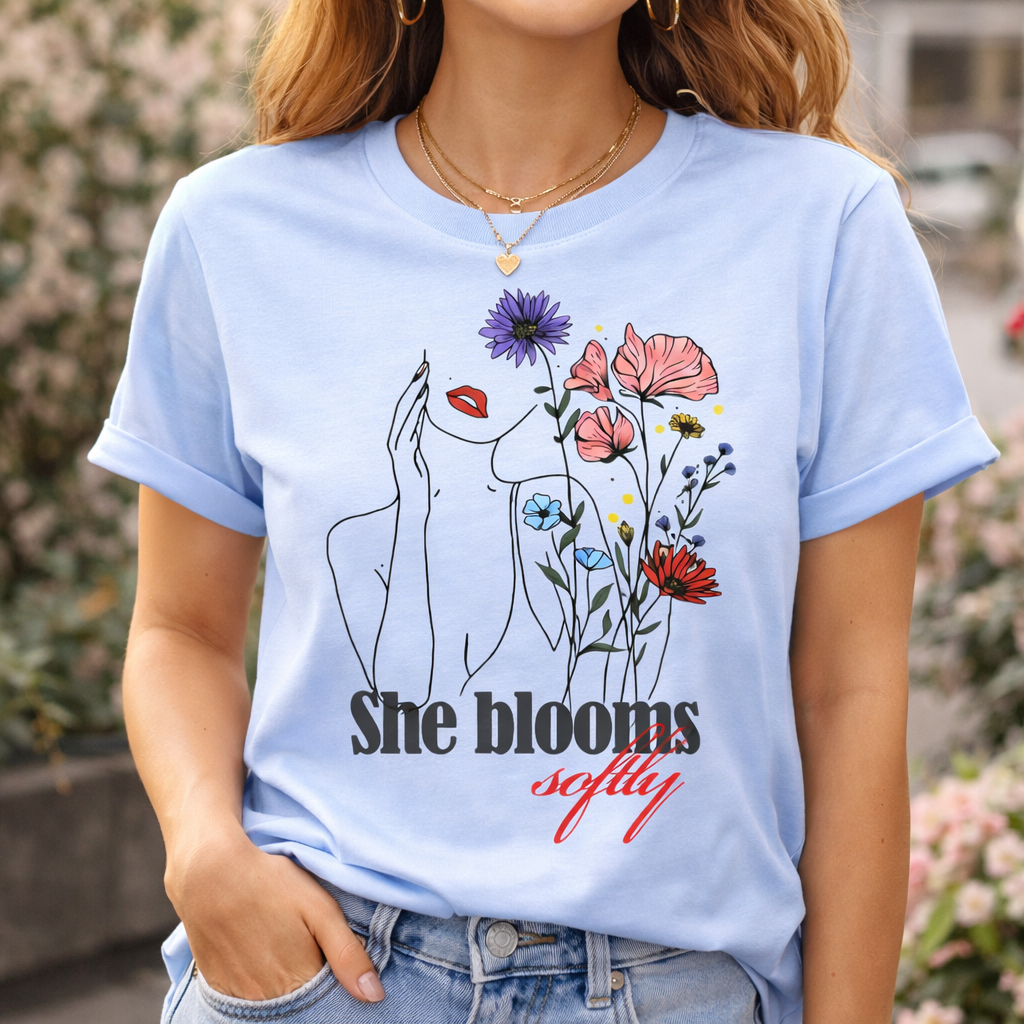 Woman posing outdoors in a light blue "She Blooms Softly" T-shirt, featuring the modern continuous line drawing and watercolor flower print.