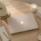 Top view of a MacBook in a beige botanical hard shell case with the Apple logo left visible, on a wooden desk with a candle, flowers and a notebook.
