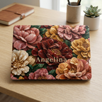 Personalized TrioAuraDesign MacBook case featuring a Stone Bouquet design with blush pink, burgundy, ivory and warm golden stone-effect flowers, deep green leaves and the custom name Angelina. The closed MacBook is displayed on a light wooden desk beside a brown notebook, pen holder and small succulent, with the Apple logo area centered in the design.