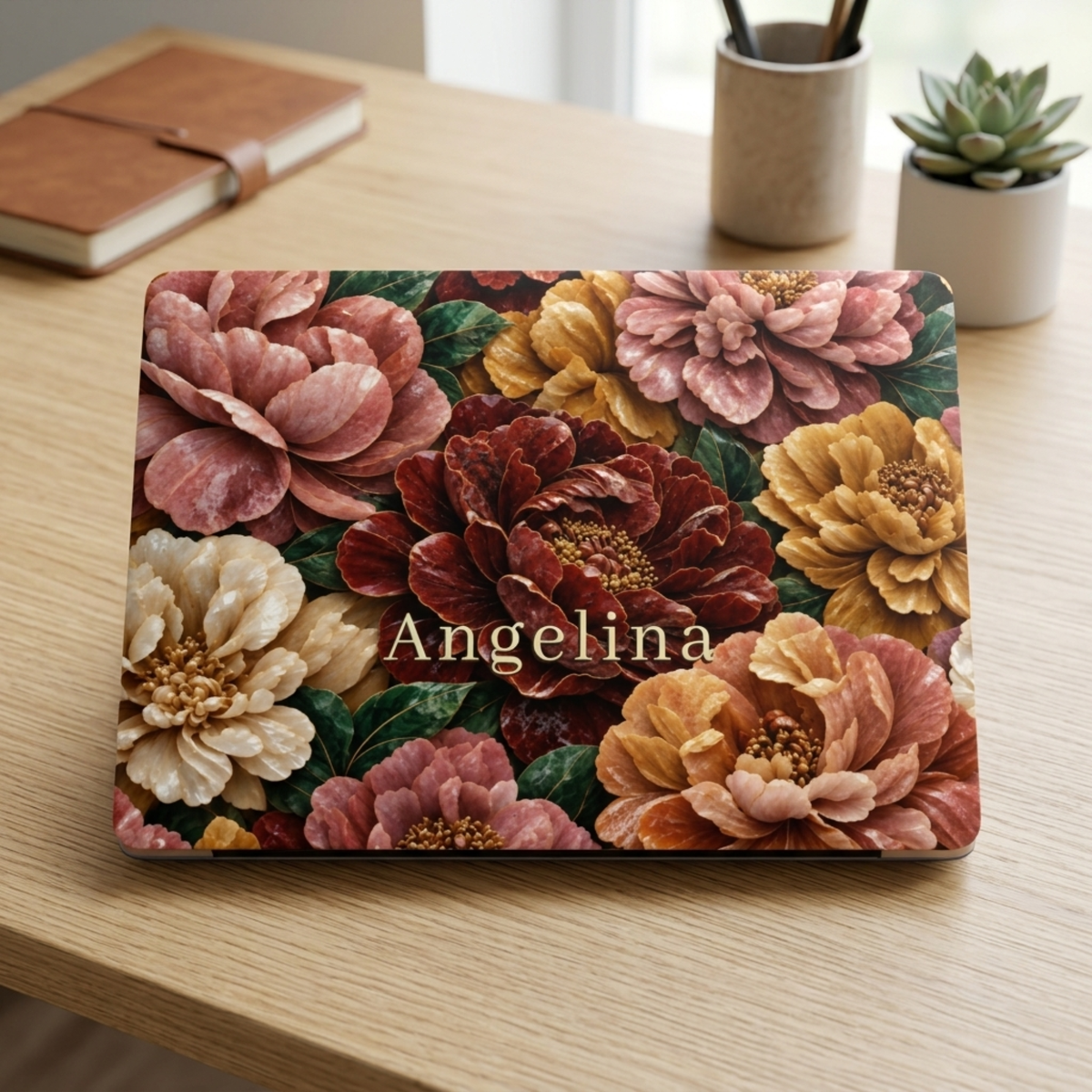 Personalized TrioAuraDesign MacBook case featuring a Stone Bouquet design with blush pink, burgundy, ivory and warm golden stone-effect flowers, deep green leaves and the custom name Angelina. The closed MacBook is displayed on a light wooden desk beside a brown notebook, pen holder and small succulent, with the Apple logo area centered in the design.