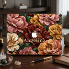 Personalized TrioAuraDesign MacBook case featuring a Stone Bouquet design with blush pink, burgundy, ivory and warm golden stone-effect flowers, deep green leaves and the custom name Angelina. The MacBook is displayed upright on a dark wooden desk beside a black and gold pen, brown notebook, coffee mug and small decorative stones, with the Apple logo visible in the center.