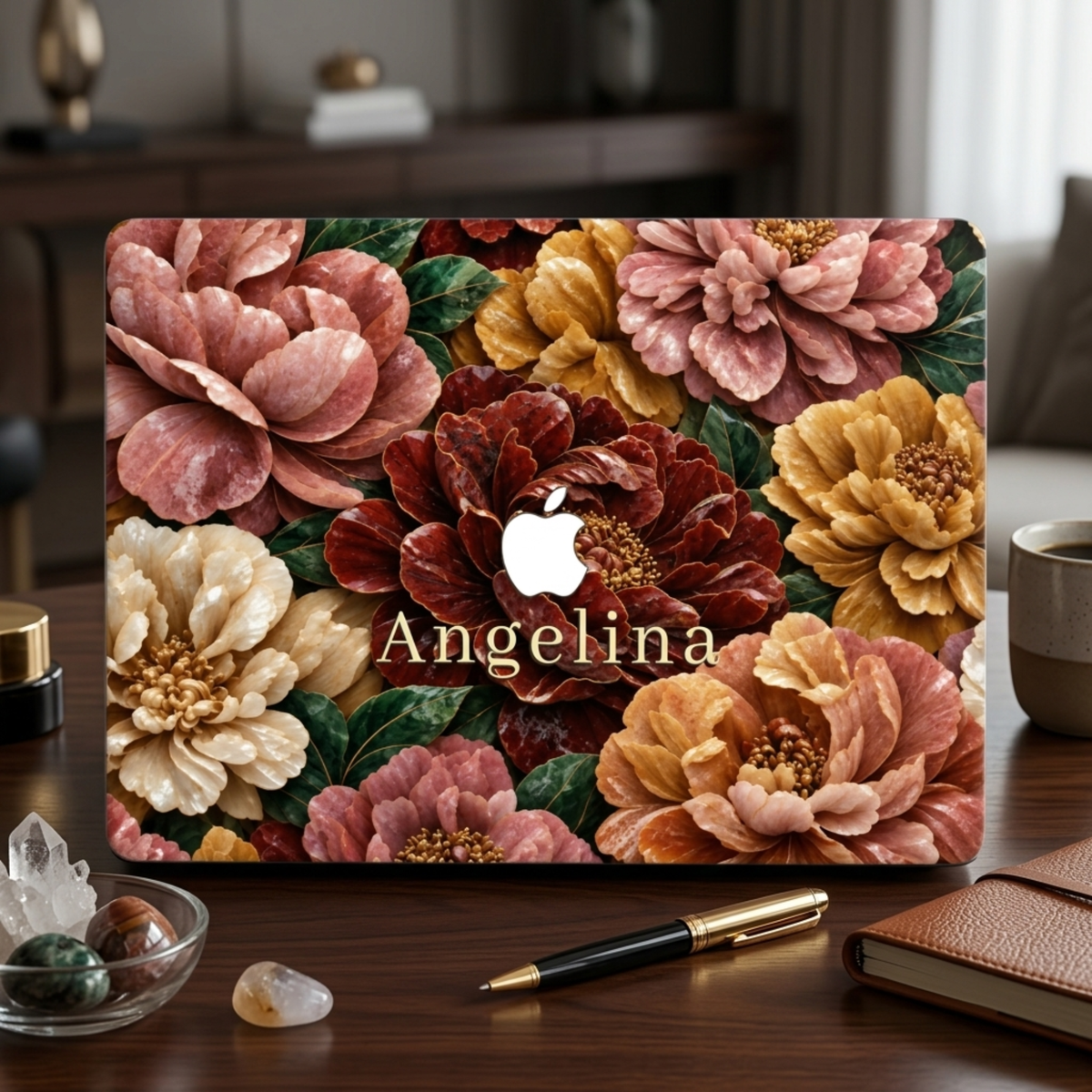 Personalized TrioAuraDesign MacBook case featuring a Stone Bouquet design with blush pink, burgundy, ivory and warm golden stone-effect flowers, deep green leaves and the custom name Angelina. The MacBook is displayed upright on a dark wooden desk beside a black and gold pen, brown notebook, coffee mug and small decorative stones, with the Apple logo visible in the center.