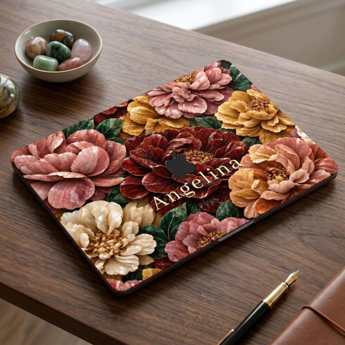 Personalized TrioAuraDesign MacBook case with a Stone Bouquet design featuring blush pink, burgundy, ivory and golden stone-effect flowers, deep green leaves and the custom name Angelina. The closed laptop is shown at an angle on a dark wooden desk beside a bowl of decorative stones, a black and gold pen and a brown leather notebook.