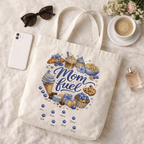 Flatlay mockup of a natural canvas "Mom Fuel" tote bag with personalized kids names, styled with sunglasses, a phone, perfume, and a coffee cup on a soft fabric background.
