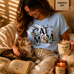 Lifestyle photo of a woman in a blue personalized 'Cat Mom' floral T-shirt relaxing with her cat, holding a 'Cat Mom Fuel' mug in a cozy room.