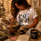Lifestyle shot of a woman working on a laptop while petting her cat, wearing a personalized 'Cat Mom' floral T-shirt with custom names Georgia and Max, in a cozy home office setting.