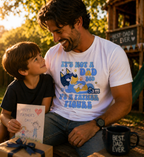 A smiling father wearing a personalized white t-shirt that says "It's Not a Dad Bod, It's a Father Figure" with a cartoon dog character, sitting outside with his young son who is holding a handmade Father's Day card.