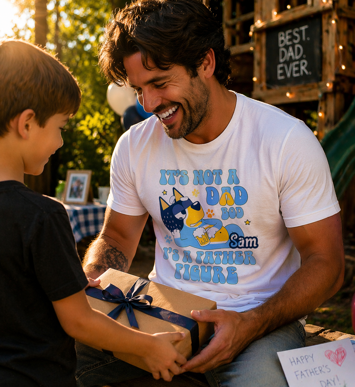 A happy father in a white personalized "Father Figure" t-shirt receiving a gift box from his son outdoors during a Father's Day celebration.