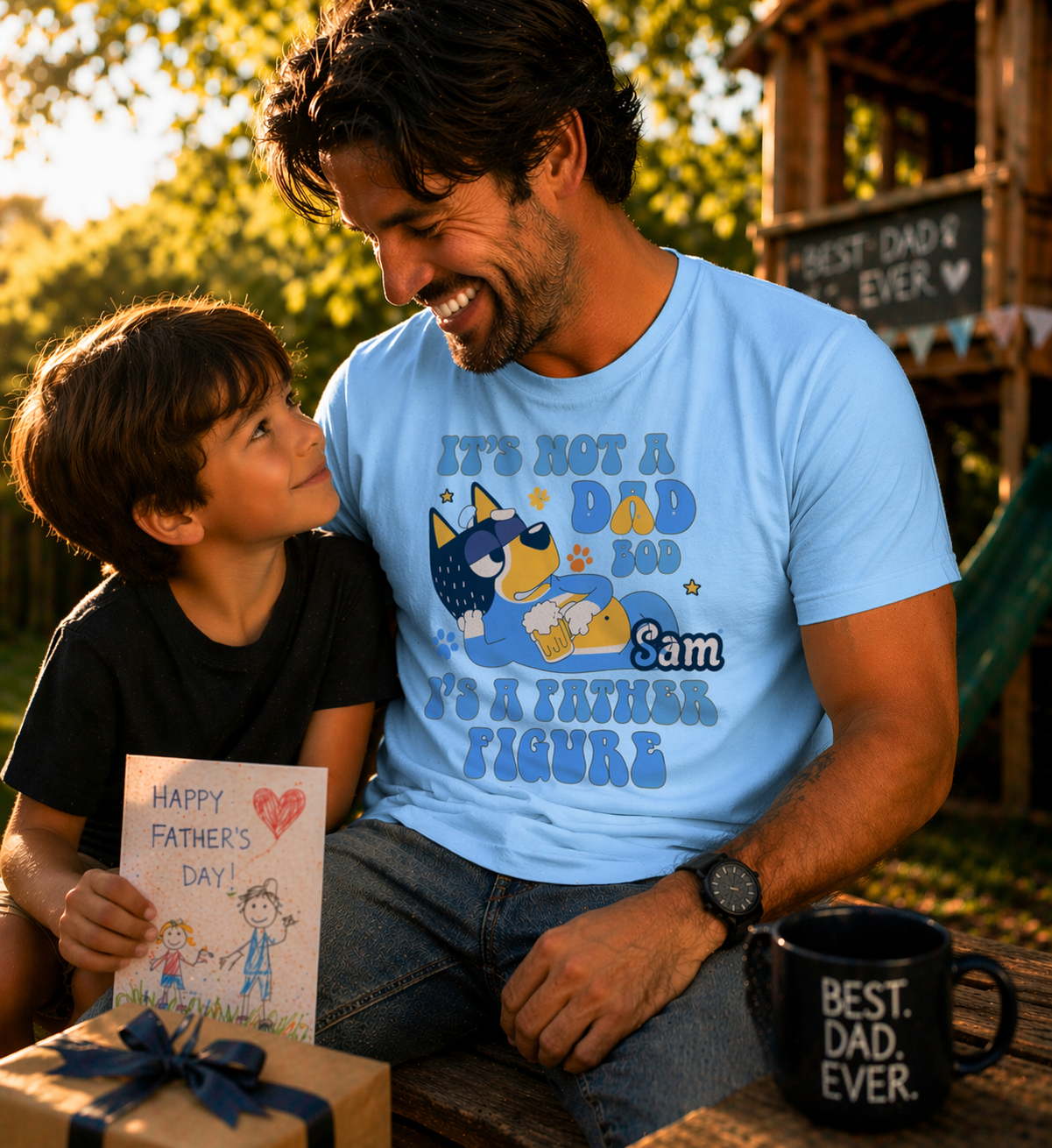 A smiling father in a light blue  personalized t-shirt with a "Father Figure" cartoon dog and the name "Sam," sitting with his son holding a Father's Day card.