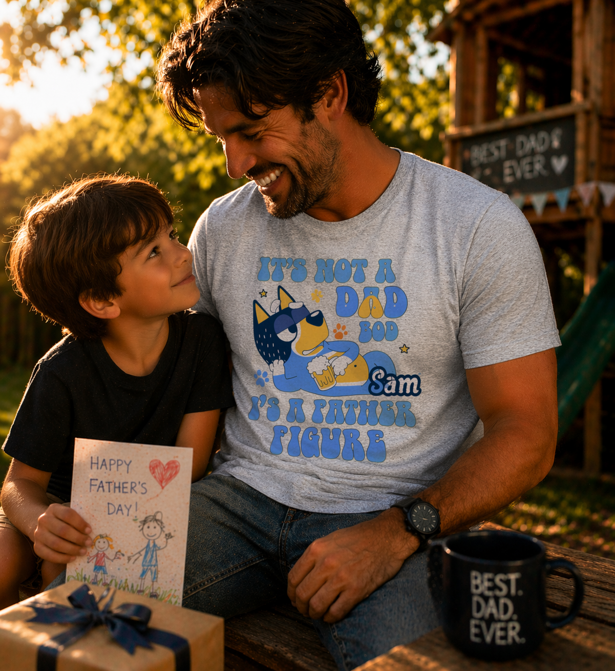A smiling father in a gray personalized t-shirt with a "Father Figure" cartoon dog and the name "Sam," sitting with his son holding a Father's Day card.