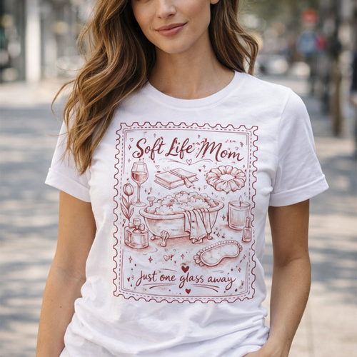 A smiling woman sitting on a beige sofa wearing a white t-shirt with a burgundy "Soft Life Mom" vintage stamp design, holding a glass of red wine. Mother's Day gift aesthetic.