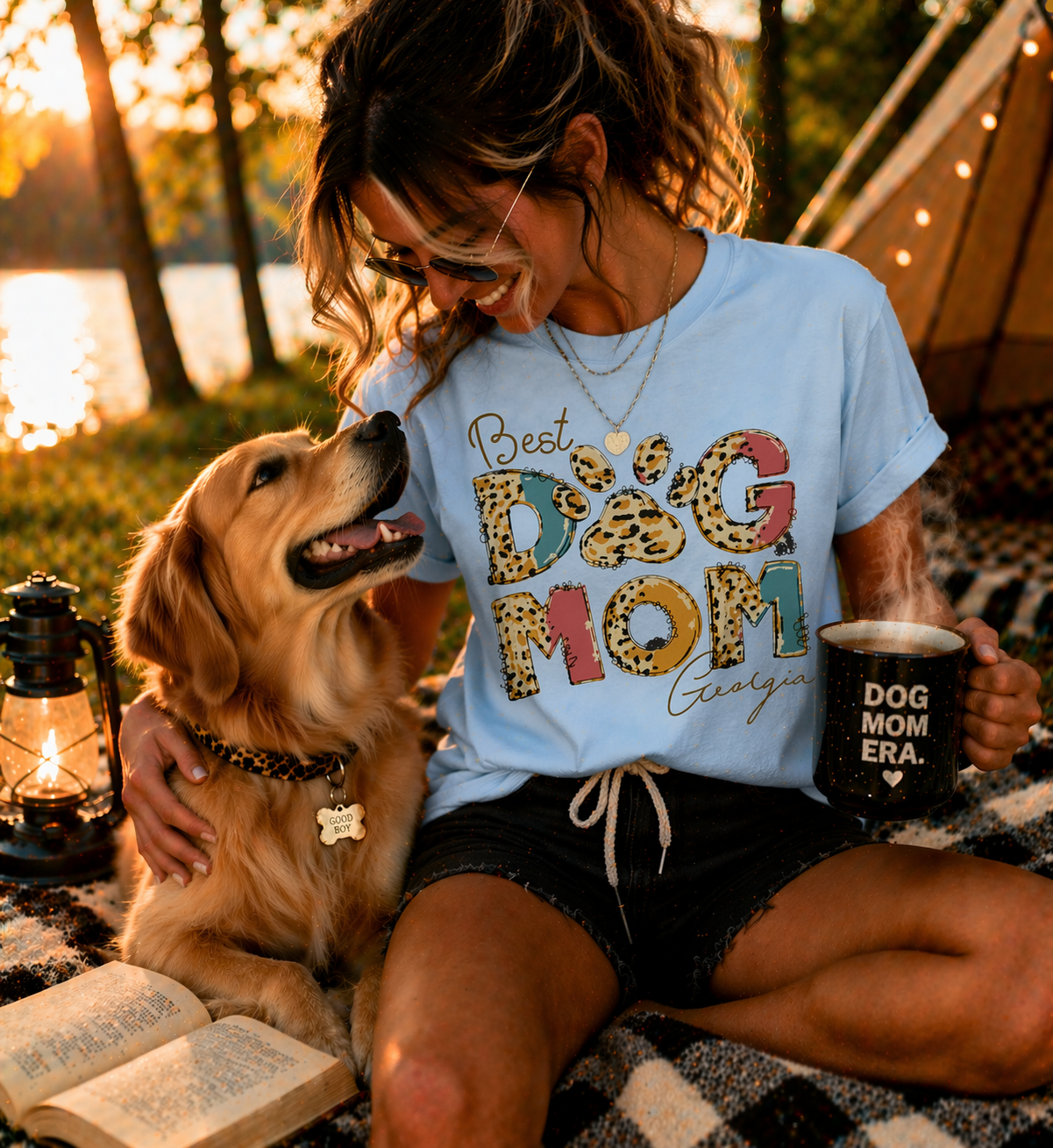 Lifestyle photo of a smiling woman sitting by a lake with her Golden Retriever at sunset, wearing a personalized light blue 'Best Dog Mom' T-shirt with trendy leopard print letters and the custom name 'Georgia' in elegant script.