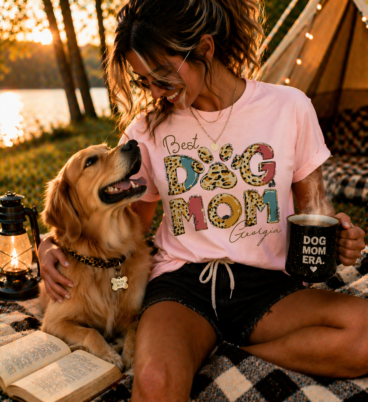 Lifestyle photo of a smiling woman sitting by a lake with her Golden Retriever at sunset, wearing a personalized pink 'Best Dog Mom' T-shirt with trendy leopard print letters and the custom name 'Georgia' in elegant script.