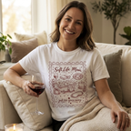 A smiling woman sitting on a beige sofa wearing a white t-shirt with a burgundy "Soft Life Mom" vintage stamp design, holding a glass of red wine. Mother's Day gift aesthetic.