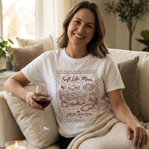 A smiling woman sitting on a beige sofa wearing a white t-shirt with a burgundy "Soft Life Mom" vintage stamp design, holding a glass of red wine. Mother's Day gift aesthetic.