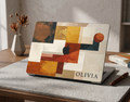A personalized abstract modern art MacBook case with terracotta, ochre, and black geometric patterns and the name Olivia, shown on a textured grey desk mat with a pen and dried flowers.