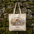 Personalized "MAMA" cotton canvas tote bag mockup with floral rainbow design and custom name Emma, hanging on a rustic mossy stone wall.