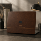 Personalized leather-effect MacBook case from TrioAuraDesign, shown on a dark modern desk with a rich brown printed leather look. The smooth hard shell features the custom text “DAD,” the phrase “ROOTED. STRONG. PRESENT.” and the Apple logo visible at center. Refined custom laptop cover with no real leather, embossing, or raised texture.