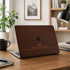 Personalized leather-effect MacBook case from TrioAuraDesign, shown on a warm wooden desk in a bright modern workspace. The dark brown smooth hard shell features the custom text “DAD,” the phrase “ROOTED. STRONG. PRESENT.” and the Apple logo visible at center. Elegant printed leather-look laptop cover with no real leather, embossing, or raised texture.