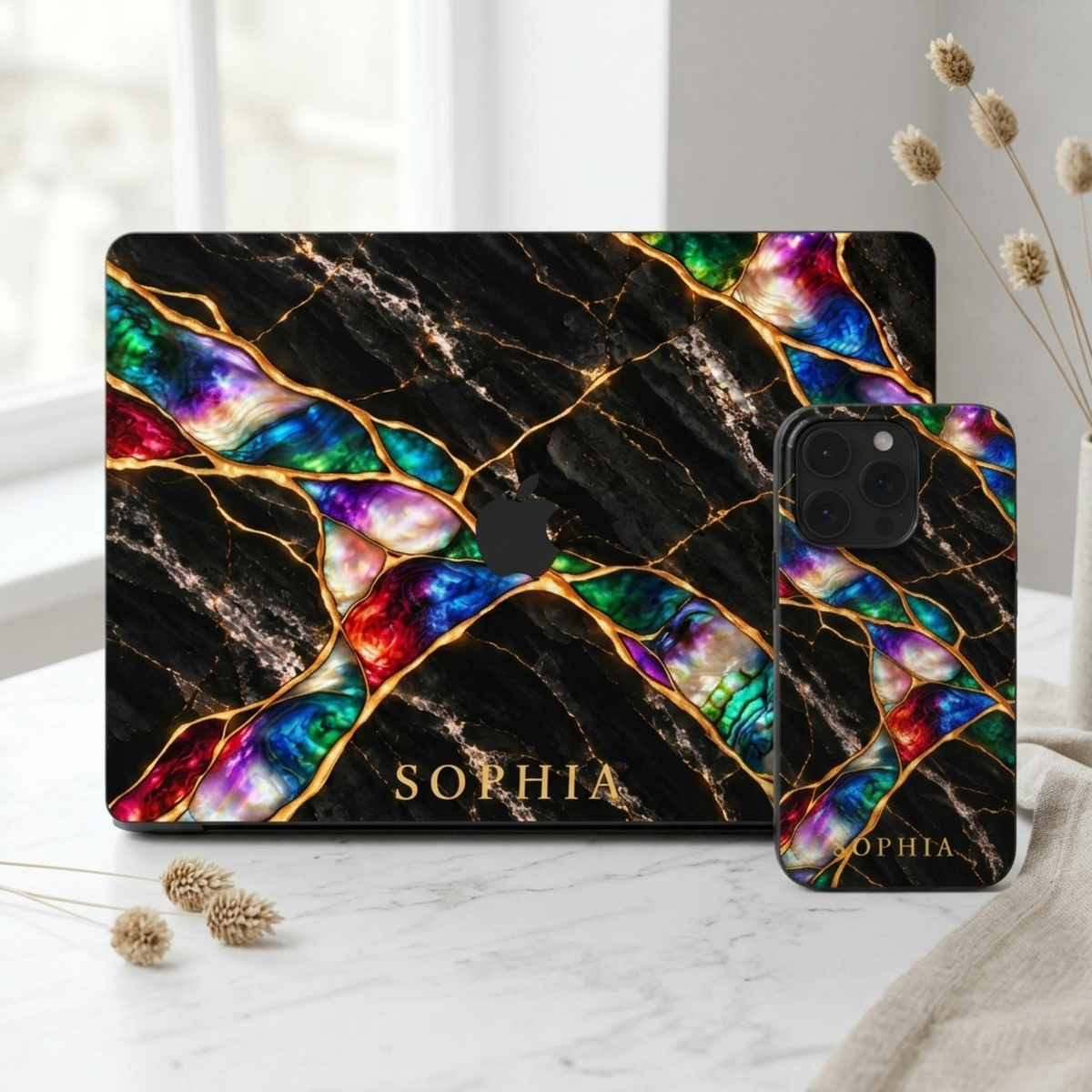 Matching set: Jewel Tone marble MacBook case and iPhone case, both personalized with SOPHIA, on a white desk.