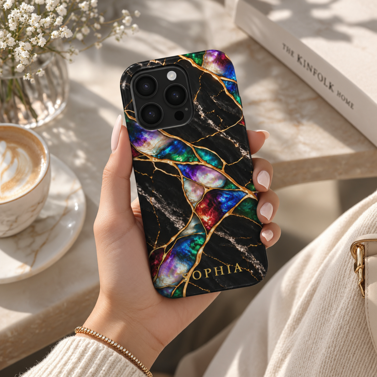 Woman's hand holding an iPhone in a Jewel Tone marble case personalized with SOPHIA, next to coffee and a book.