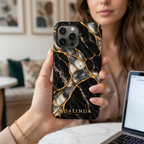 Woman holding an iPhone in a Black Pearl marble printed case personalized with ADALINDA, next to a laptop.
