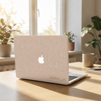 MacBook in a beige botanical hard shell case with visible Apple logo on a sunlit desk surrounded by green plants. Smooth matte printed finish.