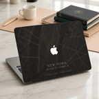 Personalized City Map MacBook case from TrioAuraDesign, shown on a light marble desk with a dark New York street map design. The hard shell features custom city text “NEW YORK,” coordinates, and the phrase “YOUR PLACE. YOUR STORY.” in warm beige lettering, with the Apple logo visible at center. Modern custom map laptop cover for compatible MacBook Air, Pro, and Neo models.