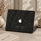 Personalized City Map MacBook case from TrioAuraDesign, shown on a neutral desk with a dark New York street map design. The hard shell features custom city text “NEW YORK,” coordinates, the phrase “YOUR PLACE. YOUR STORY,” and the Apple logo visible at center. Clean product view of a custom map laptop cover for compatible MacBook Air, Pro, and Neo models.