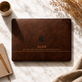 Personalized leather-effect MacBook case from TrioAuraDesign, shown on a light marble surface with a rich dark brown printed leather look. The smooth hard shell features the custom name “ALAN,” the phrase “FOCUSED MIND. KIND HEART. LIMITLESS POTENTIAL.” and the Apple logo visible at center. Elegant custom laptop cover with no real leather or raised texture.