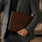 Personalized leather-effect MacBook case from TrioAuraDesign, shown being carried by a man in a gray blazer. The rich dark brown smooth hard shell features the custom text “DAD,” the phrase “ROOTED. STRONG. PRESENT.” and the Apple logo visible at center. Close lifestyle view of a printed leather-look laptop cover with no real leather, embossing, or raised texture.