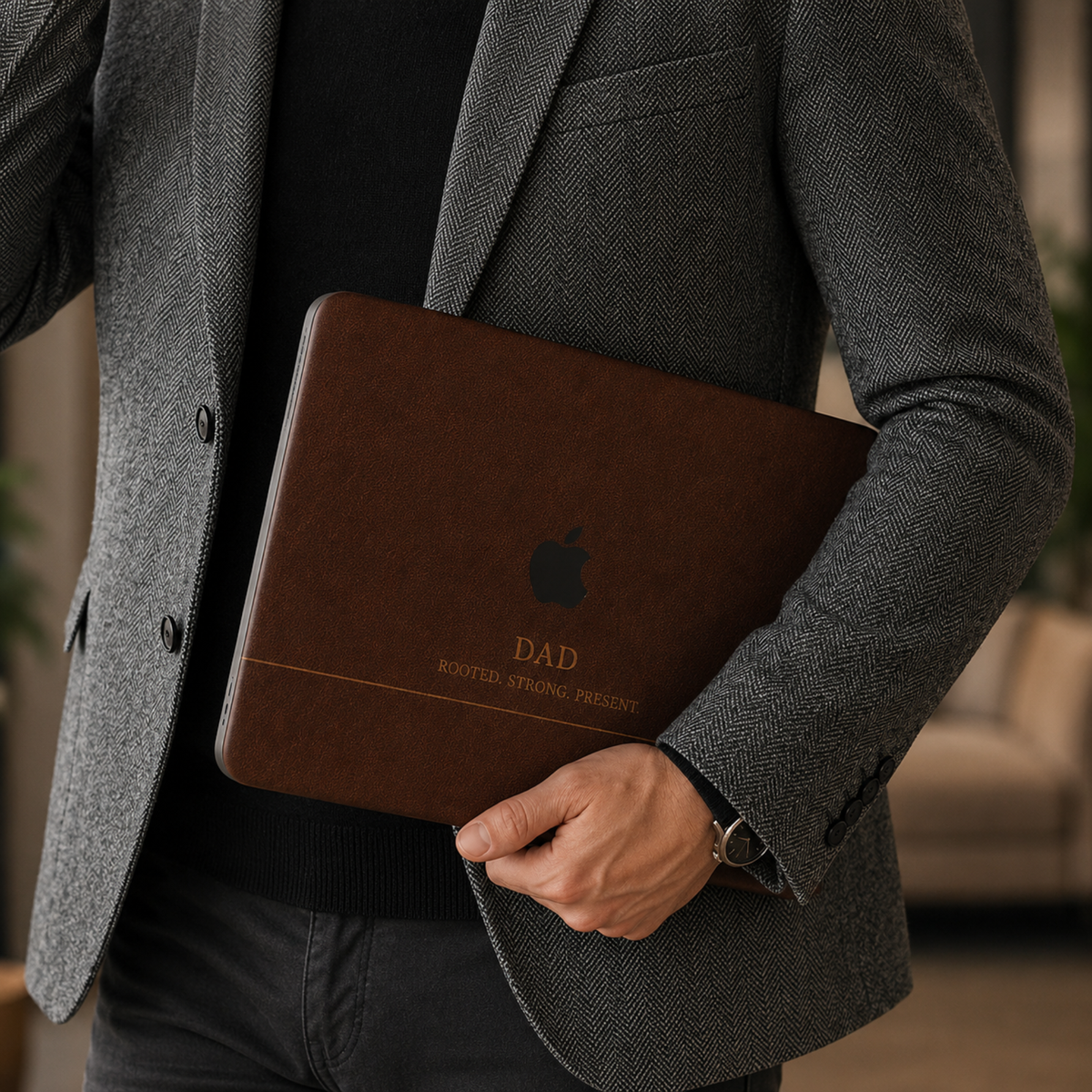Personalized leather-effect MacBook case from TrioAuraDesign, shown being carried by a man in a gray blazer. The rich dark brown smooth hard shell features the custom text “DAD,” the phrase “ROOTED. STRONG. PRESENT.” and the Apple logo visible at center. Close lifestyle view of a printed leather-look laptop cover with no real leather, embossing, or raised texture.